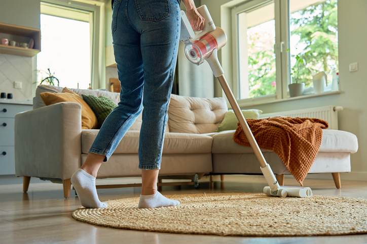 Woman is vacuuming floor of modern apartment living room, using portable cordless vacuum cleaner